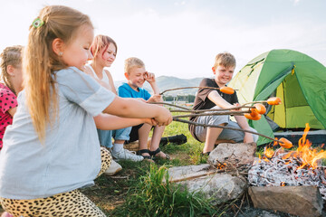 Group of Kids - Boys and girls cheerfully smiling and roasting sausages on sticks over a campfire flame near the green tent. Outdoor active time spending or camping in Nature concept.
