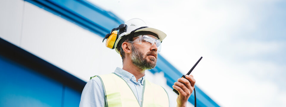 Man engineer wearing a helmet and protective glasses talk on walkie-talkie. Bearded engineer on produce site. Wide image