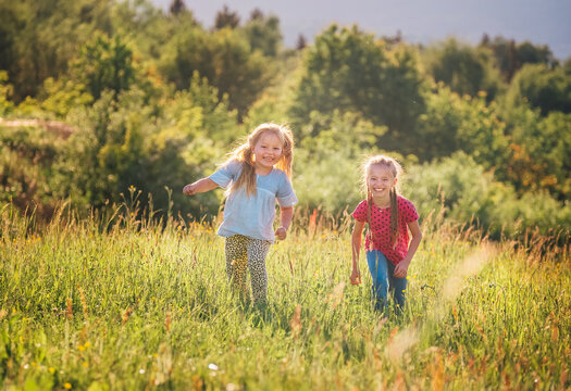 Two little sisters running and laughing in the summer high green grass meadow with a bright sunset light background. Careless childhood, family values, and outdoor activities concept photo.