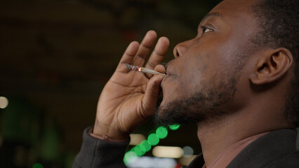 Close-up young handsome African American man smoking cigarette in dark alley. Lips blowing smoke on...