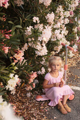 Child girl in pink summer dress sitting on a pink kick scooter near bushes of oleander nerium in bloom flowers of pink and white colors - 510940264