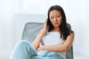 Confused upset tanned lovely young Asian woman listen shocked news at call using phone at home...
