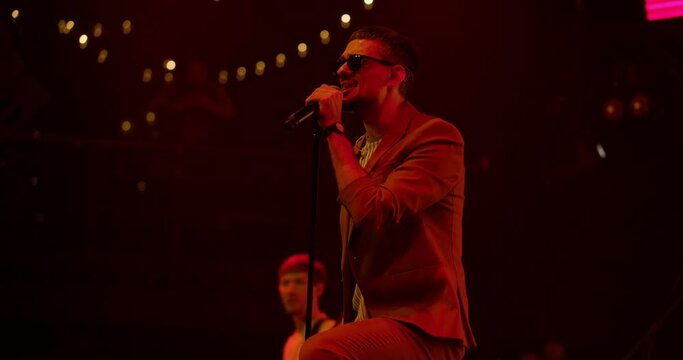 Portrait of singer man in glasses performing on stage at concert. Singer singing into microphone. Live music party in front of bright colorful strobing Lights