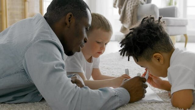 Loving family African American father and son together with caucasian child draw on floor in living room. Black man and his son decorate drawings together with caucasian boy lying on floor of house.