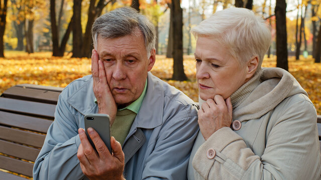 Upset elderly married couple sit on bench in autumn park read bad news on smartphone worried old people look at screen phone sad family feeling shocked read unpleasant message internet fraud concept