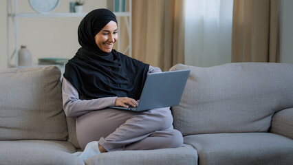 Friendly arabian muslim woman girl in hijab sit on cozy sofa talk video call with friend greeting...