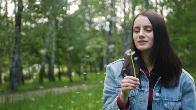Charming caucasian beauty in jeans jacket blowing on a dandelion on a sunny summer day. Cute brown-haired woman blowing seeds of dandelion seadhead and smiling childishly and genuinely to the camera