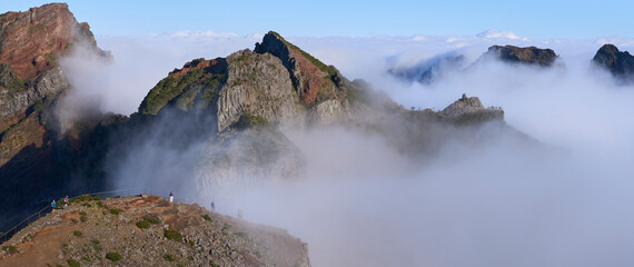 Panorama from Pico do Areeiro, a starting point of PR1 trail to Pico Ruivo. Fog ascending from a valley and remaining among mountain folds. Mountains slopes with mist. Tourists on a hiking trail.