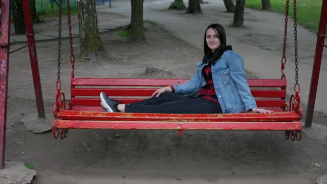 Cute brown-haired caucasian woman in jeans jacket swaying on a big swing. Attractive smiling female swinging and relaxing in park. Cheerful girl laying comfortably and rocking slowly on a swing 
