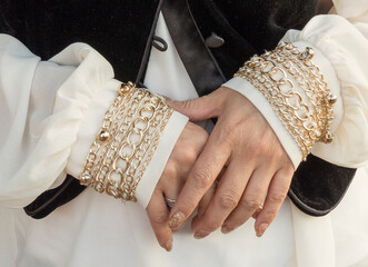 closeup of woman's hands with manicured nails and sleeves ending in cuffs that have golden chains sewn onto them, she's wearing a velveteen vest with satin trim