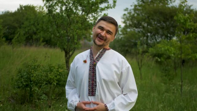 Handsome caucasian man wearing a beautiful white vyshyvanka outdoors. Cheerful young slav dude in traditional embroidered Ukrainian and Belarussian shirt smiling and enjoying nature in grassy meadow

