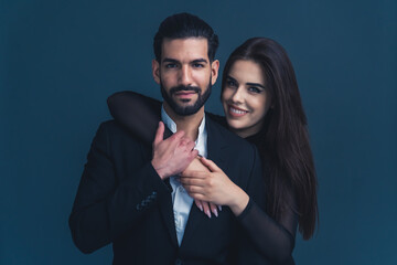 Studio shot of a professional heterosexual couple in formalwear. Bearded dark-haired Cuban young male entrepreneur being hugged bu his long-haired girlfriend with a beautiful smile. High quality photo