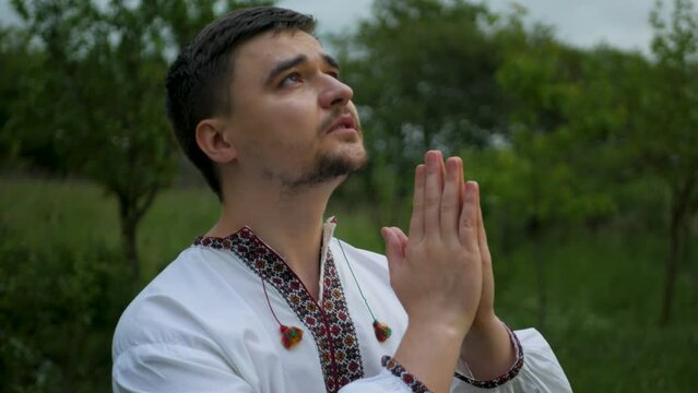 Hopeful and calm caucasian man in traditional Ukrainian national outfit folding hands and praying to God for everything to be alright. Religious person's in embroidered vyshyvanka prayer for peace
