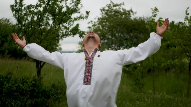Adult caucasian male wearing Ukrainian traditional shirt spreading hands to sides and looking up outdoors. Man in embroidered vyshyvanka looking to the sky and talking to God with open arms in nature
