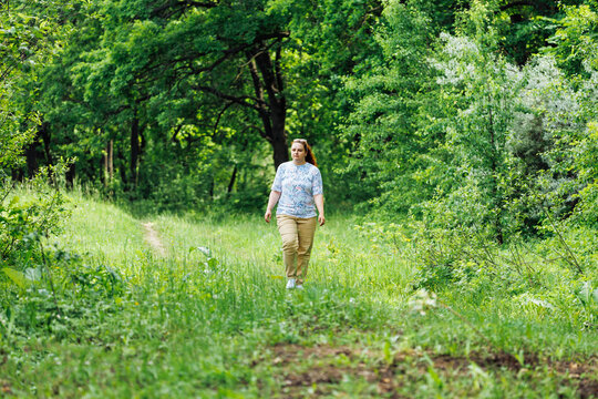 Portrait of young buxom woman with long curly red hair in ponytail, wearing blue blouse with floral pattern, beige trousers, sunglasses walking in park forest among green trees bushes. Summer, nature.