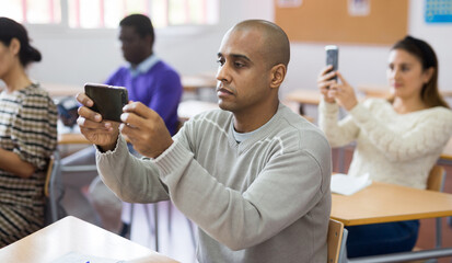 Portrait of focused hispanic man using smartphone to record learning material during lecture in school for adults