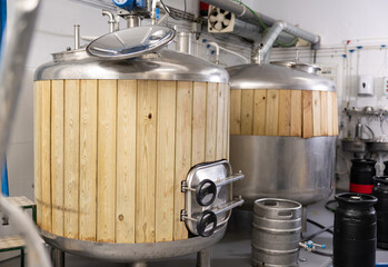 Cylindrical steel fermenters with wood trim and kegs in brewery shop. Craft brewing system. Beer...