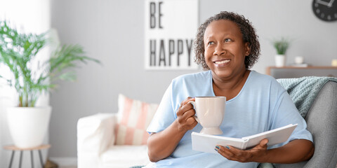 Happy mature African-American woman with cup of tea and book at home. Concept of ageing and...