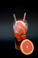 Grapefruit juice in a tall glass with ice and straws on a black background, next to pieces of ripe citrus.