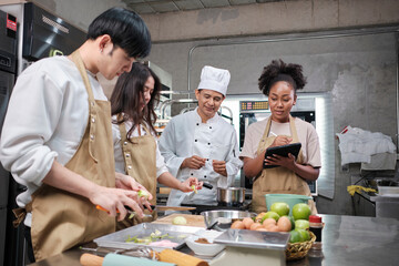 Hobby cuisine course, senior male chef in cook uniform teaches young cooking class students to peel and chop apples, ingredients for pastry foods, fruit pies in restaurant stainless steel kitchen.