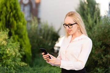 Portrait of a beautiful joyful business woman in a shirt and black skirt, using her smartphone, standing against the background of a flower bed
