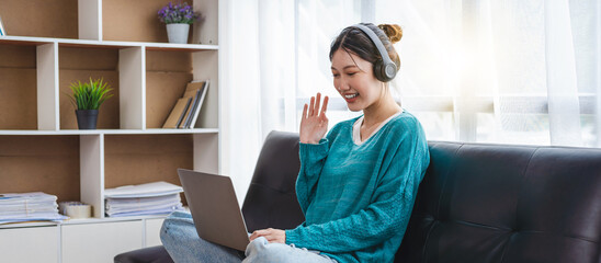 Attractive asian woman sitting on sofa, relaxing and talking to her friend or family, waving at the laptop screen. - obrazy, fototapety, plakaty