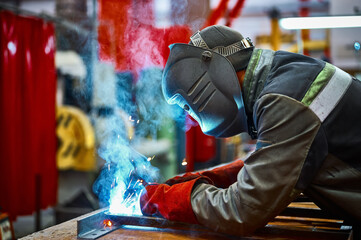 Skilled worker in protective mask welds metal part at plant