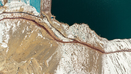 Aerial view of the reservoir of the plaster and its dam stopping the turquoise water. dirt road with tourists walking and touring the Andean sector in a winter landscape with snow in the mountains