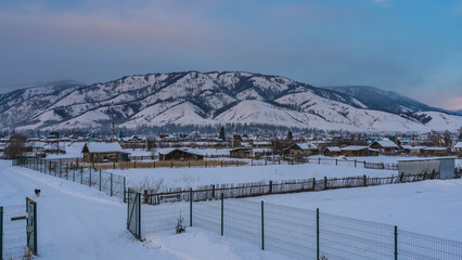 Fototapeta premium Siberian village in a snowy valley. One-story wooden houses, fences are visible. A picturesque mountain against the blue sky. Altai