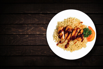 Top view of instant noodles in white bowl with added pieces of chicken on dark wooden background.