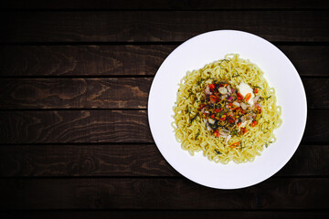 Top view of instant noodles in white bowl with added garlic and chili on dark wooden background.