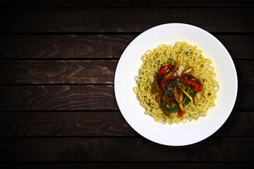 Top view of instant noodles in white bowl with added chopped vegetables and black peppers on dark wooden background.