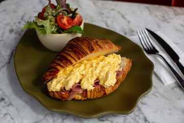 Delicious and healthy breakfast - croissant with ham, eggs, and salad from tomatoes, lettuce on green plate.