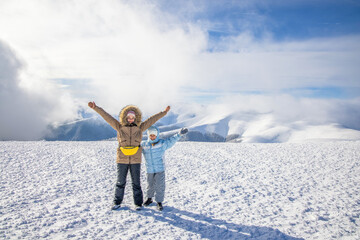 Fototapeta na wymiar Two girls in winter clothes pose spreading their arms on a mountain against the backdrop of the cloud-covered sun