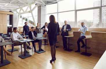 Fototapeta na wymiar Group of diverse business people listen to speech by female speaker at business meeting. Experienced business lady speaks in front of audience in loft office. Concept of business speeches.