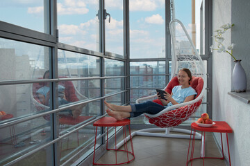 Barefoot pretty woman in blue t-shirt and jeans sits on hanging lounge seat and uses tablet. Relaxing rocking wicker white chair with red cushion on home or hotel loggia with panoramic windows. - obrazy, fototapety, plakaty