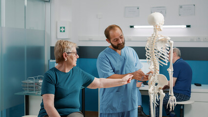 Osteopath explaining orthopedic diagnosis on human skeleton to elder woman at recovery checkup....