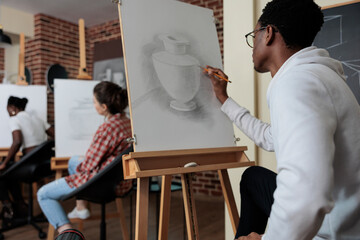 Creative student sitting in front of white canvas drawing vase model using graphic pencil working at sketch illustration during art class. Diverse team attenting sketching lesson in creativity studio
