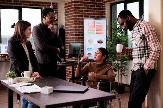 Asian woman wheelchair user with disability laughing with people in startup office, meeting on break. Female worker dealing with chronic health condition, talking to cheerful colleagues.