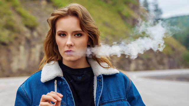 young woman in a denim jacket smokes a cigarette near the road.