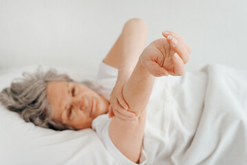Fototapeta na wymiar Rested senior woman stretching after sleeping in bedroom in morning. Waking up mature woman doing health exercises while relaxing in bed. Selective focus on female hands, close-up