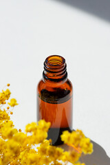Open amber bottle without cap with serum or essential oil with yellow flowers. White background with daylight and beautiful shadows. Beauty concept for face and body care