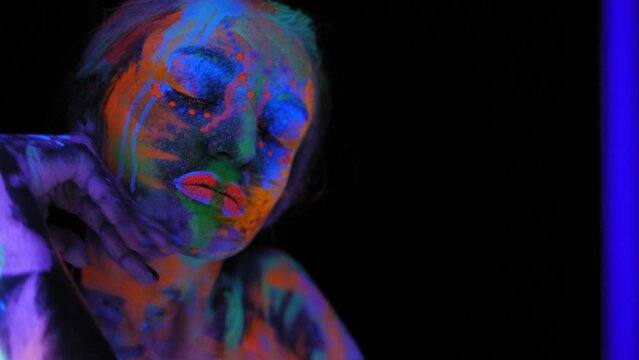A magical space girl with UV drawings on her face and body in the dark, she moves her hands near her face. Body art.