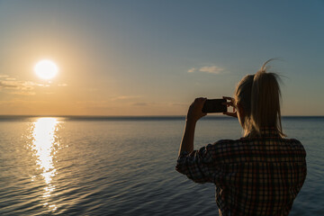 A young attractive woman takes a photo of a sunset over a river with her mobile phone. View from the back of a girl stopping to take a photo or enjoy a beautiful view while traveling