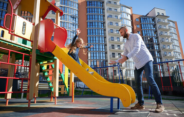 Handsome dad catching cheerful daughter, riding on slide at modern courtyard of city residential...