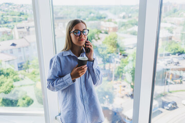 Beautiful young business woman talking on smartphone and working on new productivity project for company, spending free time in coworking center enjoying window view