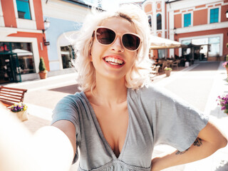 Young beautiful smiling hipster woman in trendy summer clothes. Sexy carefree model posing on the street background at sunset. Positive blond female outdoors. Cheerful and happy. Taking Pov selfie