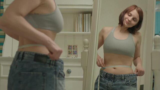 30s woman measuring her waistline with measuring tape looking at the mirror. Happy woman after weight loss.