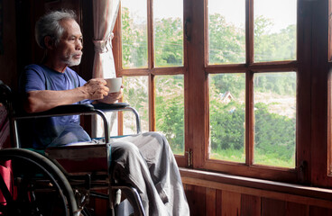 A senior Asian man in a wheelchair holds a cup of coffee and looks at the window.