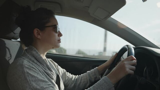 Young woman with sunglasses driving a private car on the road during sunny day. Profile side shot of female driving on highway or country road. Relaxed enjoying the way as buildings and trees pass by